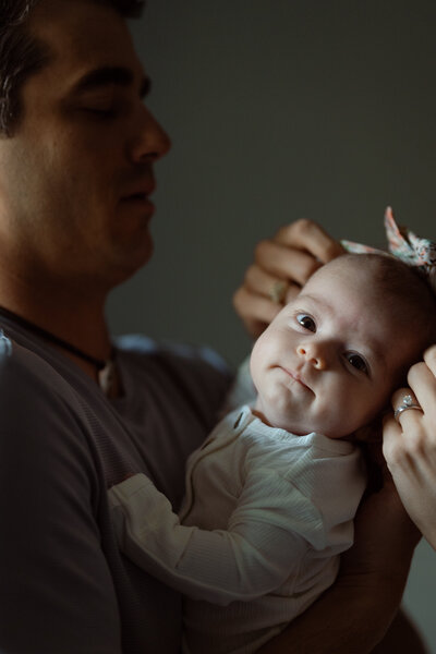 A newborn looks gently into the camera while being held by her father, as her mother adjusts a delicate headband. Captured by JPF Galleries, this lifestyle moment is softly lit and full of warmth.