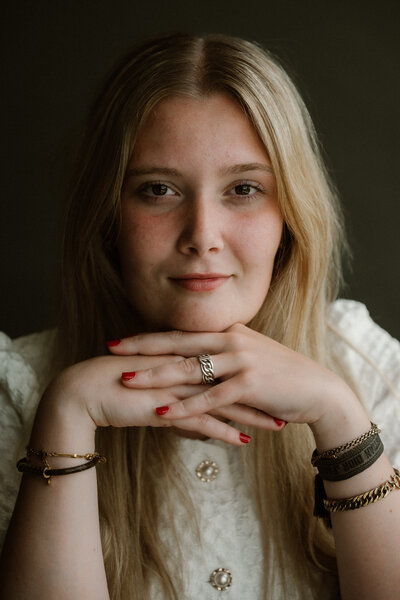 Portretfoto jonge vrouw met haar handen gevouwen onder haar kin in een studio setting met daglicht.