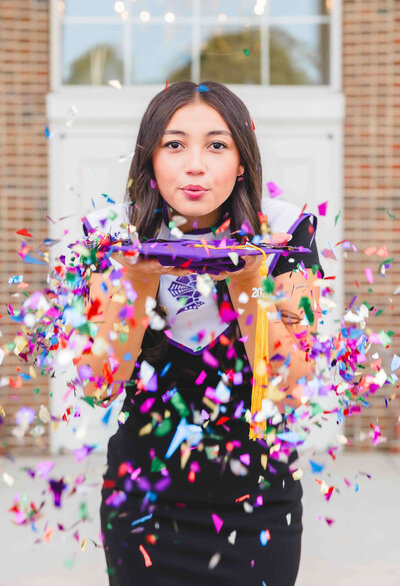 A person in a black outfit stands proudly in front of a building, holding a graduation cap with a tassel. As colorful confetti flies towards the camera, the scene—captured brilliantly by a senior photographer in Fayetteville, NC—features a brick wall and white door in the background.