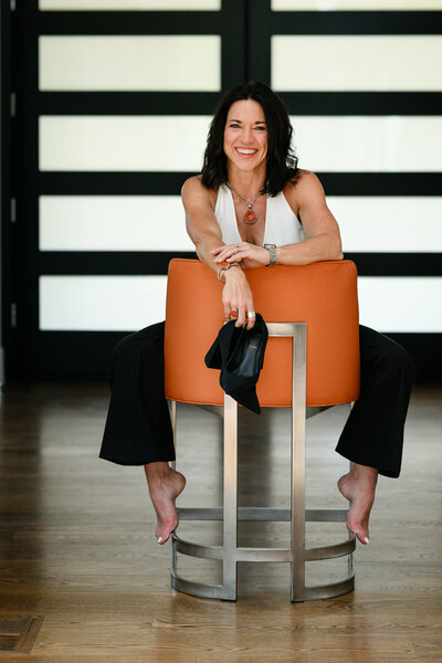 Denver branding photographer captures a woman business owner sitting backwards in an orange chair holding her high heels for her marketing photos for her small business.