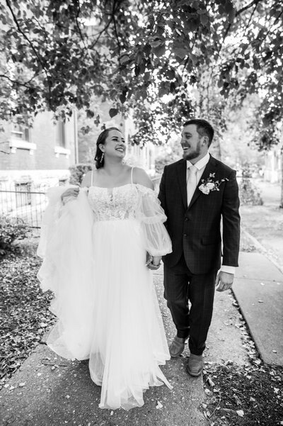 Bride in a white dress walking down the sidewalk with her groom dressed in a black suit
