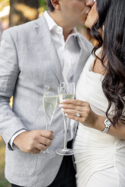 A couple kisses while holding champagne glasses. The man is wearing a light gray suit, and the woman is in a white dress with an engagement ring and bracelet. Her long dark hair is styled in loose waves.