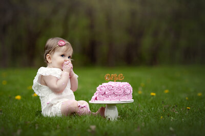 One year old girl sitting in grass eating a pink smash cake.