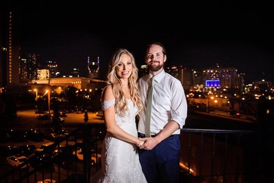 Night photo of bride and groom standing on the balcony of their wedding venue after sunset with the Nashville skyline lit up behind them