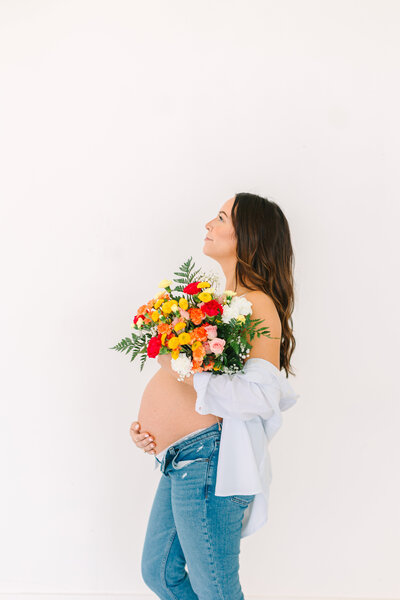 Pregnant woman in jeans with a shirt hanging off her arms holding flowers over her pregnant belly in front of a white wall