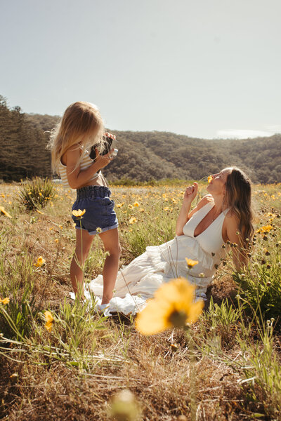 Woman in a white dress sitting in a field of wildflowers, laughing, while a young girl in shorts and a tank top stands taking a photo of her with a camera. Hills are visible in the background.