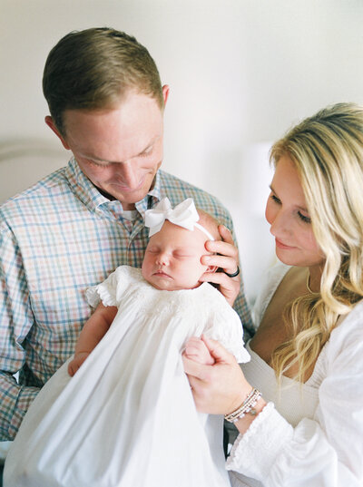 Father holding newborn daughter in white dress and bow upright while mother holds her hand and they both look lovingly at her taken by Little Rock Photographer Bailey Feeler