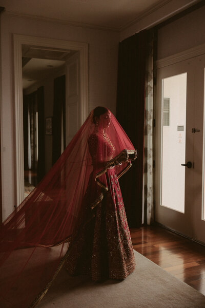 South Asian bride wearing a red bridal lehenga with a long veil, standing in soft indoor light before her ceremony.