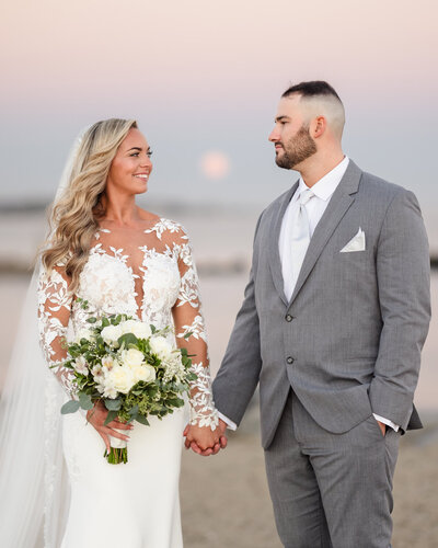 Bride and groom holding hands at sunset, gazing at each other with love and calm joy.