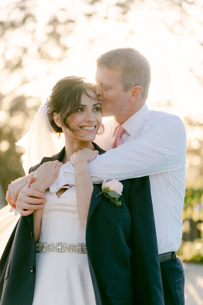 bride wearing groom's jacket as he hugs her from behind and kisses her on the forehead during golden hour romantics at el encanto a belmond hotel in santa barbara, ca captured by magnolia west photography los angeles wedding photographers