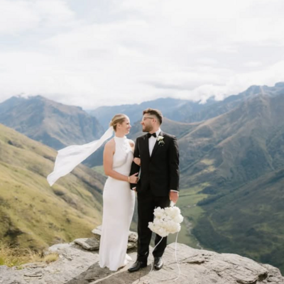 Bride and groom standing on a rocky mountain ledge, with the bride's veil flowing in the wind and dramatic mountain scenery in the background. Melissa Clark Photography