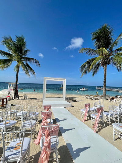 A beautifully decorated beachfront wedding ceremony setup with white chairs, a pastel aisle, and an ocean view, planned by a destination wedding travel agent in Mexico.