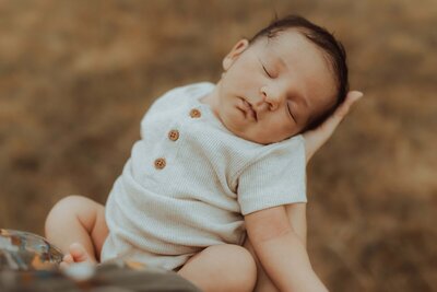 A baby wearing a cream-colored onesie with wooden buttons is sleeping peacefully while being held in someone's hand. 