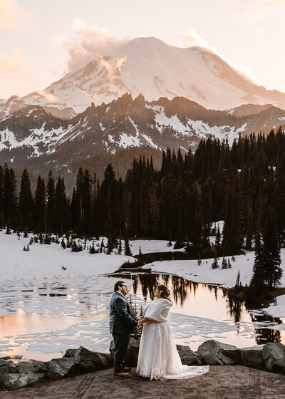 A couple in wedding attire stands above an alpine lake covered in snow. a snowy mountain top rises from behind the towering pines. Pink, golden light paints the highlights of this image. We're offered a unique perspective as their adventure elopement photographers shoot down from a higher vantage point. 