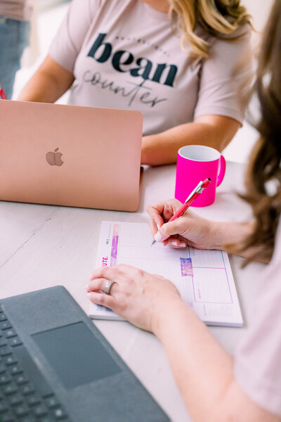 Personal Brand Image of woman writing notes on a weekly planner with another woman behind her typing on a rose gold Apple laptop