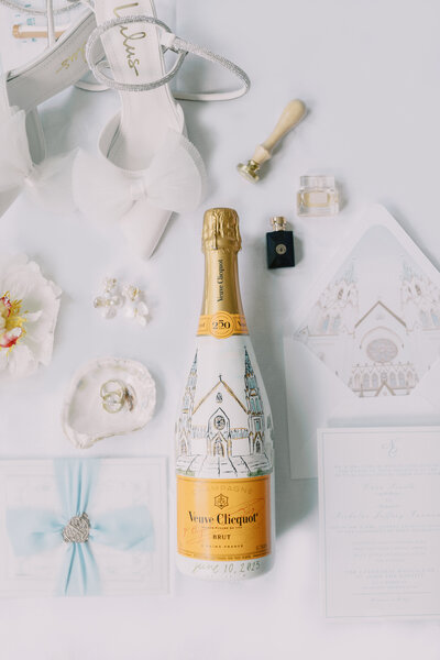 Image of selection of bridal details on white background including orange customized and painted champagne bottle with venue painted on the side. The bottle is surrounded by an open oyster with bridal rings, and invitation pieces. Also around the bottle are white bridal heels with chiffon bows on the toe and oyster earrings, small perfume bottle and wax press.