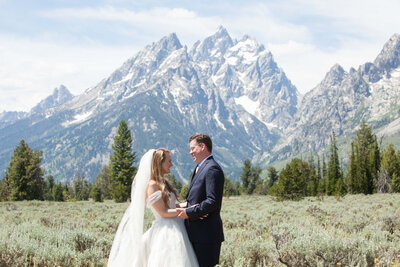 A bride and groom share a moment at Grand Teton National Park, standing in a lush green field with jagged mountains in the background.