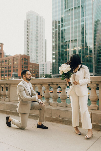 Man in tan suit kneeling and proposing to woman in white pantsuit holding white flowers in downtown Chicago