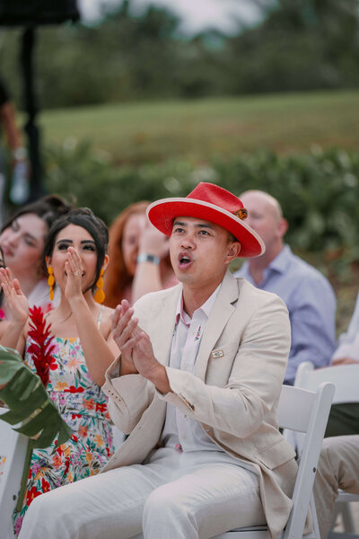 During a beach wedding ceremony in Mexico, a man wearing a red hat enthusiastically claps.