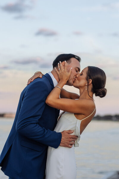 Romantic moment as the bride and groom share a kiss at sunset, set against a serene waterfront backdrop at Crystal Point Yacht Club. The bride, in a sleek white gown, embraces the groom in a navy suit, with soft pastel hues of the evening sky reflecting their love.