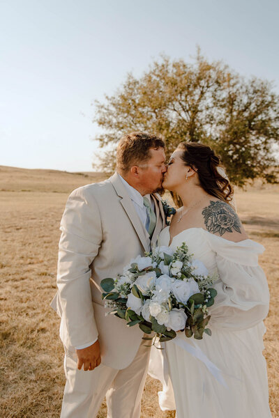 Bride and groom kissing in a golden field with bouquet in hand