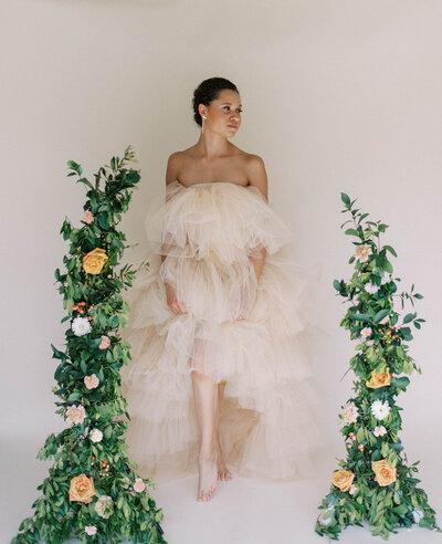 bride in a light beige tulle gown that is off the shoulder stand in between two tall greenery and floral decorations