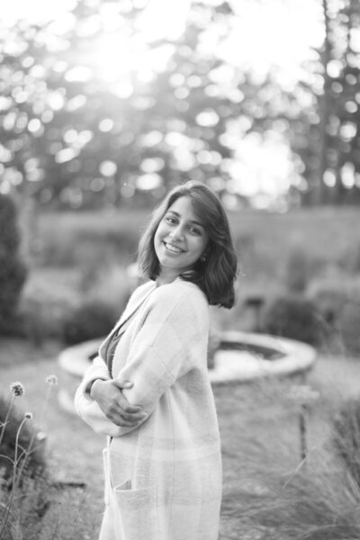 A woman stands outdoors, smiling with arms crossed. She wears a light cardigan and has shoulder-length hair. The background features blurred foliage and sunlight filtering through trees, creating a serene, natural setting. The photo is in black and white.
