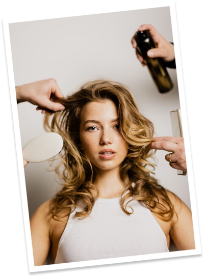 A blonde female with a blowout with hands brushing and using hairspray during a photoshoot for social media.