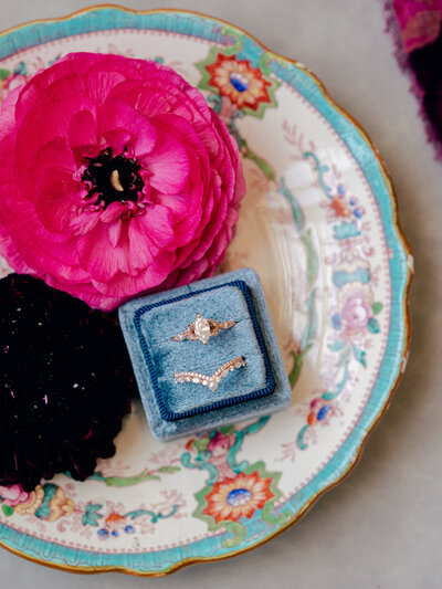 An engagement ring in a blue ring box sits in an antique floral patterned dish. Flowers surround the ring box.