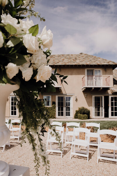 Floral decoration with white roses in foreground, white chairs lined up and house in background
