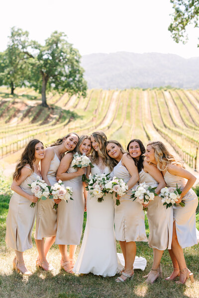 bride with her bridesmaids dressed in champagne silk dresses and hugging in a napa vineyard.