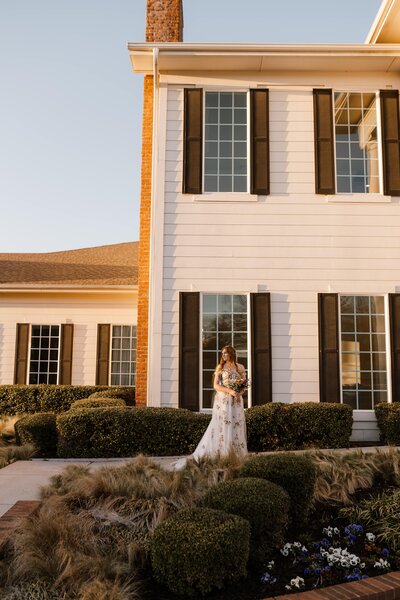 Bride standing in front of her wedding venue, The Milestone Denton, looking to her right into the sunset holding her bouquet. Capture by Shelly Voss
