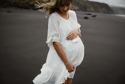 A close up portrait of a mother to be holding her belly and wearing a white dress during her beach maternity photoshoot in new plymouth