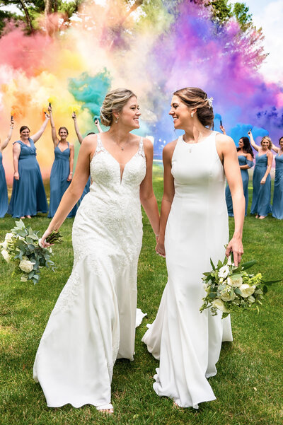 Two brides holding hands and walking in front of rainbow smoke bombs. The venue is now called the Legacy Club but was formally known as Woodcrest Country Club Cherry Hill NJ 