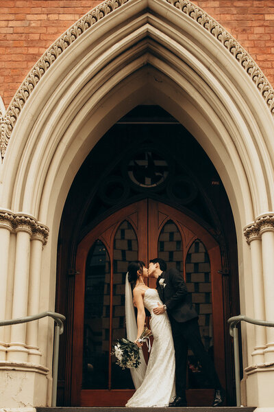 Bride and groom share a kiss under the grand arched entrance of a cathedral on the Sunshine Coast.
