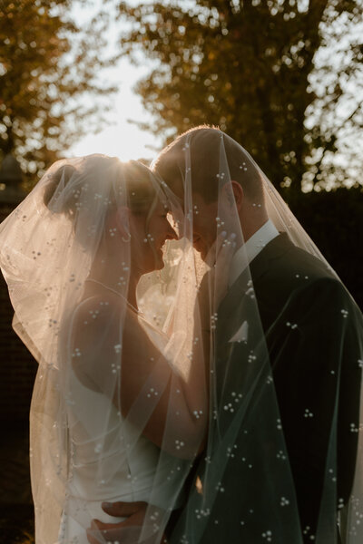 A bride and groom stand closely, enveloped in a softly lit veil under dappled sunlight. Their foreheads touch, creating a tender, romantic atmosphere.