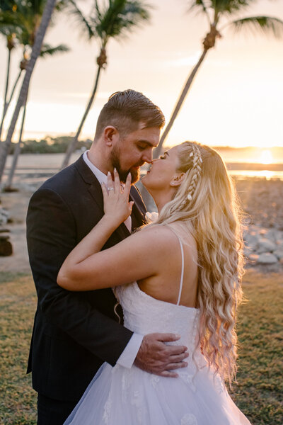 Bride and groom sharing an intimate kiss with the sun setting behind them, while the bride softly touches the grooms cheek while kissing.