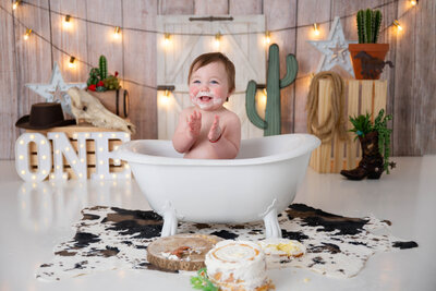 Smiling baby clapping while sitting in a mini tub, surrounded by cowboy-themed props including barrels, cow hides, and cactuses.