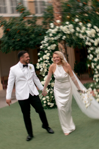 photo of a bride and groom holding hands while smiling and running towards the camera away from a white floral wedding arch