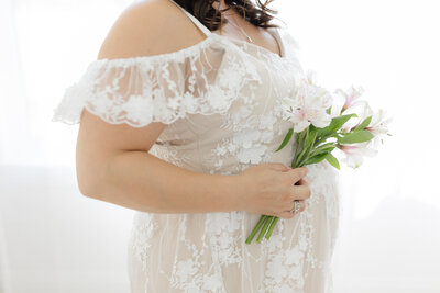 close up of pregnant mothers belly wearing white lace dress and holding flowers