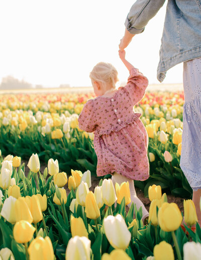 A little girl in a pink dress holds her mother's hand behind her as she runs through yellow tulip fields.