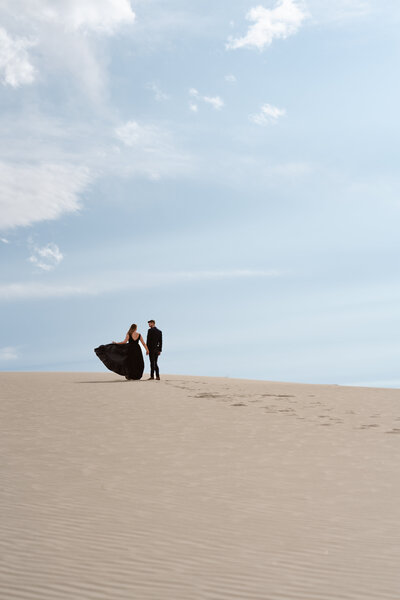 A couple in black clothes stands on top of a sand dune silhouetted against the sky