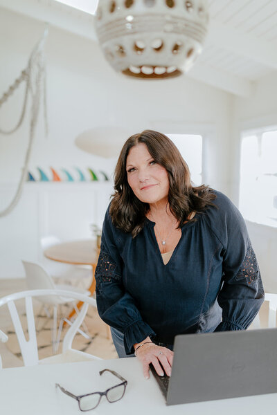 Susie Schumacher standing at a desk with a laptop in front of her, smiling softly in a modern white office with ocean-inspired decor - Susie Schumacher Life Coach