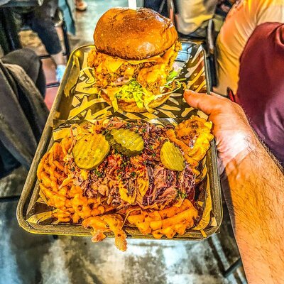 A tray with a fried chicken sandwich on a brioche bun, paired with curly fries topped with pulled pork, coleslaw, and pickles.