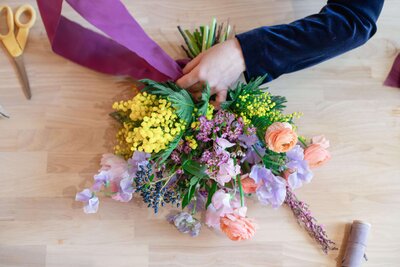 Close-up of two hands tying a purple ribbon around a colorful spring bouquet with mimosa, ranunculus, sweet peas, and lilac flowers on a light wood table.