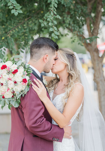 A bride and groom kiss on their wedding day. The groom is wearing a maroon tux. The bride has her bouquet around him as she hugs and kisses him.