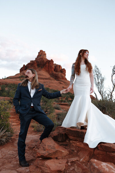Elopement couple at Bell Rock in Sedona, Arizona—standing together and squared to the camera, holding one hand between them while looking in opposite directions. She stands slightly higher on the red rock, creating a bold, editorial feel against the stunning desert backdrop