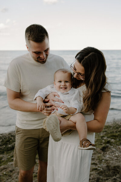 Sweet family photography session by Morgan Ashley Lynn Photography on the beach at Lake Michigan in Milwaukee, WI with mom holding toddler boy and looking down at him, dad has his arms around son too looking down and smiling, toddler boy is looking straight on to camera smiling wide