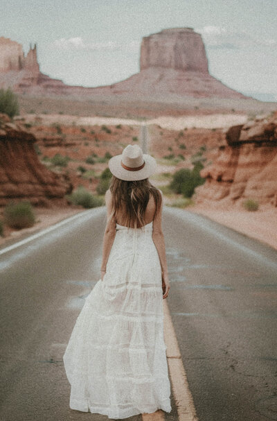 A woman in a white dress and hat stands on a deserted road in a desert landscape with large rock formations, capturing the spirit of adventure that inspires Sweet Pea Ink Creative's approach to brand design.