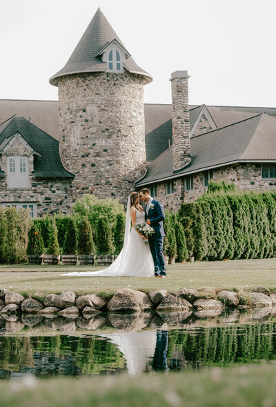 A bride and groom stands near a pond at a castle with a reflection in the water